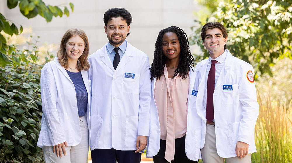 Four medical students wearing white coats standing outside on campus